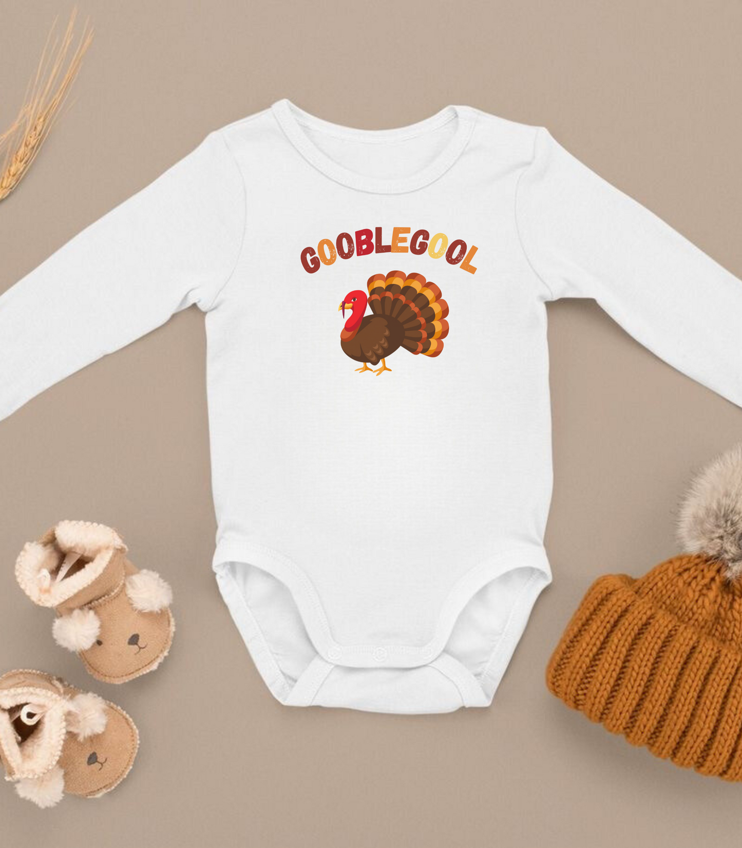 A white long sleeve bodysuit for babies with 'GOOBLEGOOL' printed in red and green colors, accompanied by a brown hat and tan boots.