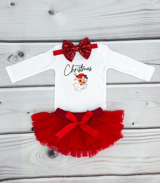 A white infant jumpsuit with a 'Merry Christmas' text design, red bow detail, and a small red Santa hat, presented on a wooden background.