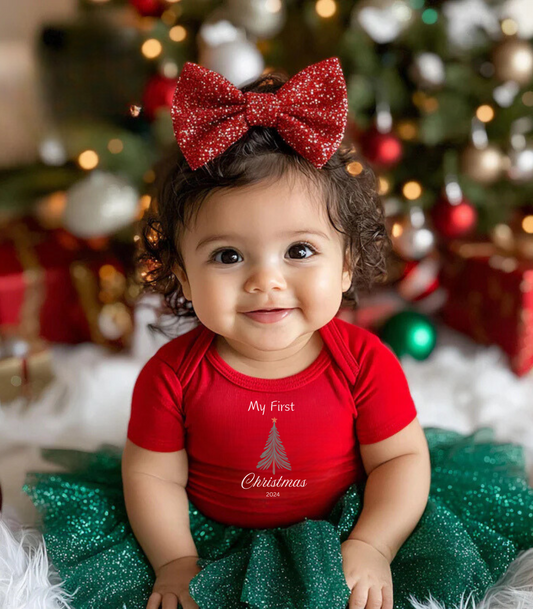A baby wearing a red Christmas-themed jumpsuit with a bow and a Christmas tree on the front, surrounded by holiday decorations.