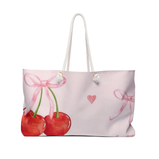 A tote bag with a pink background featuring a large cherry design in the center, accompanied by smaller cherries and hearts. The bag has white rope handles.