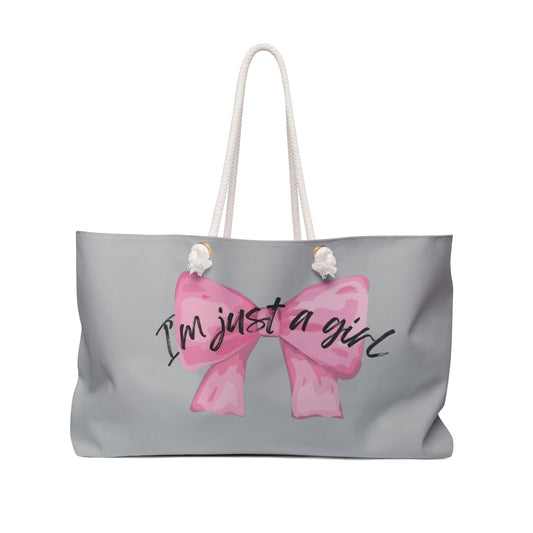 A large tote-style weekender bag with a gray background featuring a floral print. The bag has the text 'I'm just a girl' accompanied by a pink bow graphic on the front.