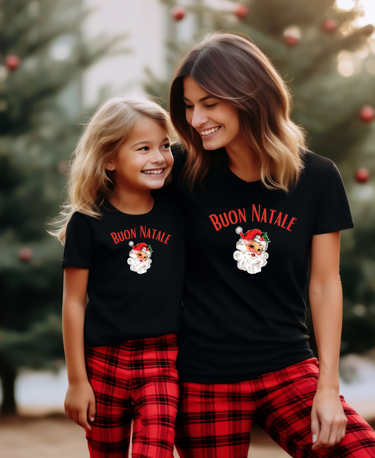 An image showing two individuals, an adult and a child, wearing matching black Christmas t-shirts with 'Buon Natale' and 'Merry Christmas' text in red and white colors, standing near a Christmas tree.