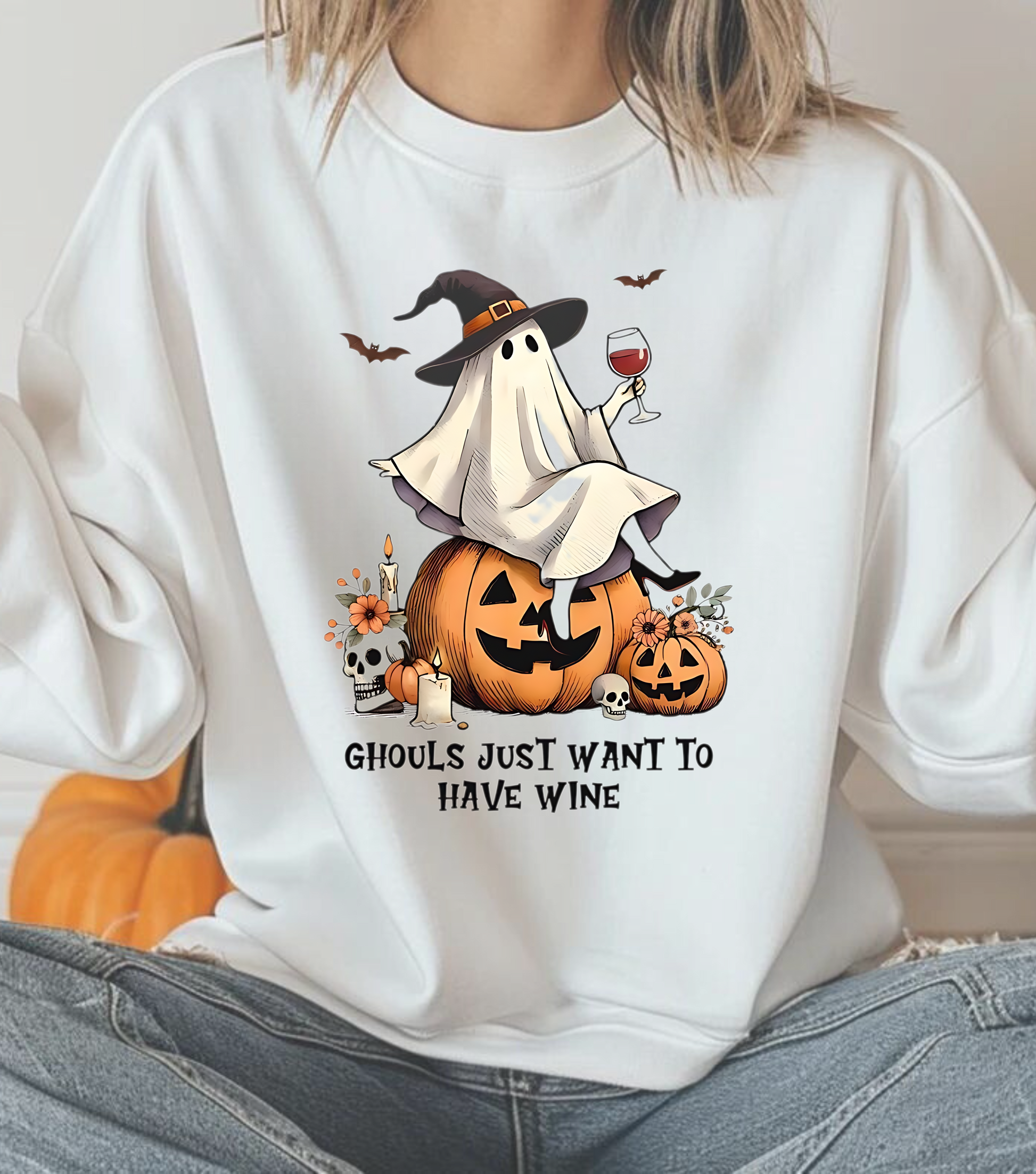 A person wearing a white crewneck sweatshirt with a graphic of a ghost, a pumpkin, and a glass of wine, along with the text 'Ghosts just want to have wine'.