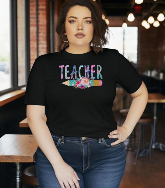 A person wearing a black short-sleeve t-shirt with the text 'Teacher' and a graphic of a pencil and flowers on the front, standing in a room with a brick wall and wooden furniture.