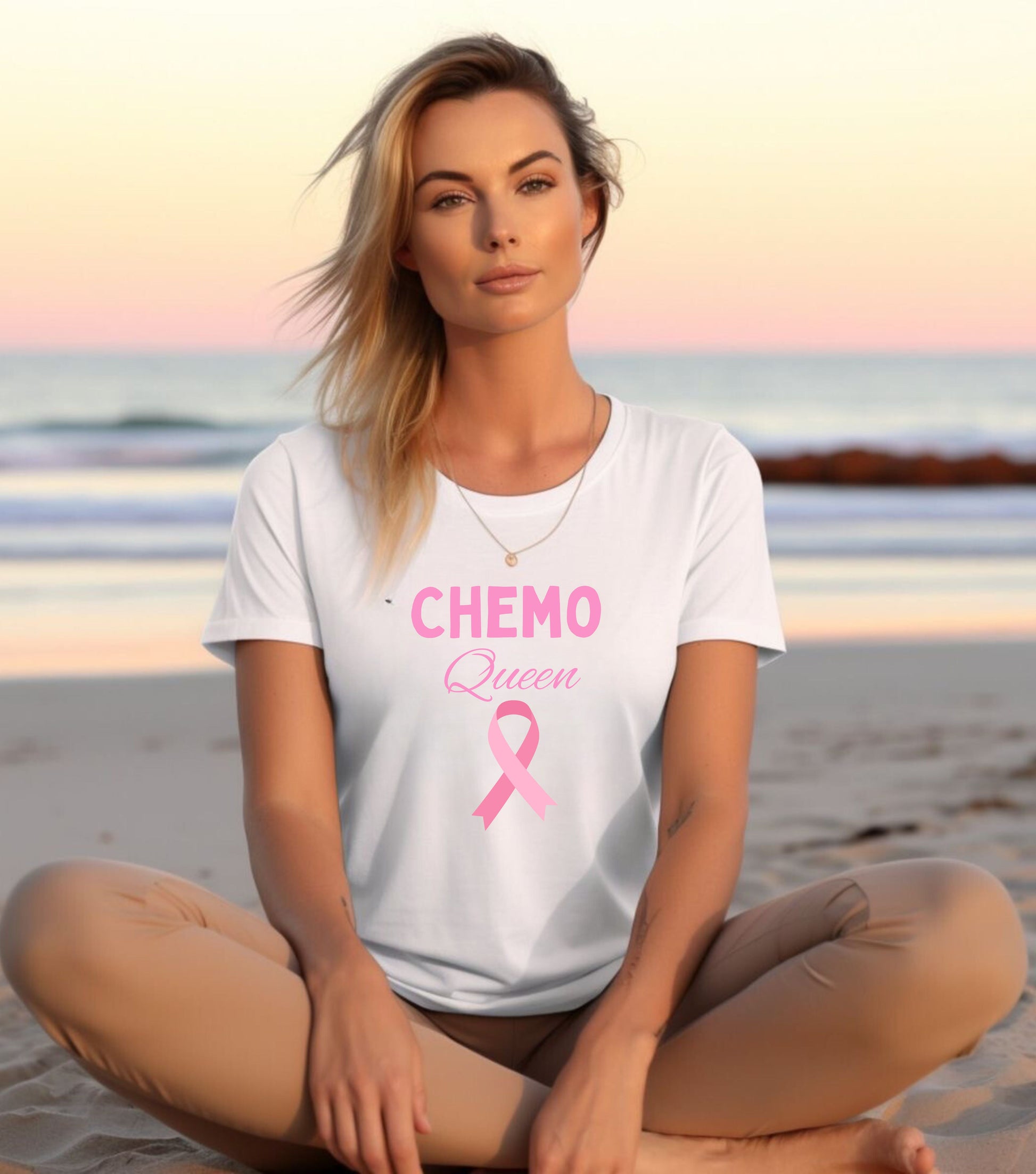 A person sitting on a beach wearing a white unisex short-sleeve t-shirt with the text 'CHEMO QUEEN' and a pink ribbon symbol on it, indicating breast cancer awareness.