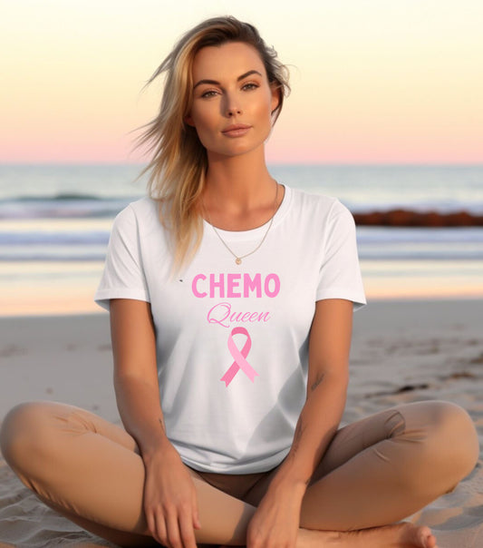 A person sitting on a beach wearing a white unisex short-sleeve t-shirt with the text 'CHEMO QUEEN' and a pink ribbon symbol on it, indicating breast cancer awareness.