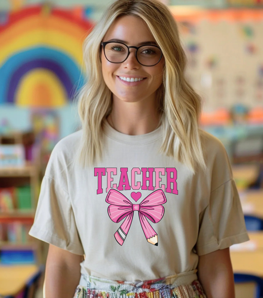 Woman wearing a 'Teacher' shirt with a pink bow graphic in a classroom setting